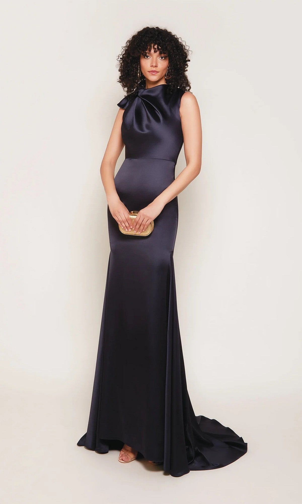 A woman wearing the Alyce 7067 long white sleeveless satin gown with a sheath silhouette and bow neckline holds a small gold clutch, standing against a plain background.